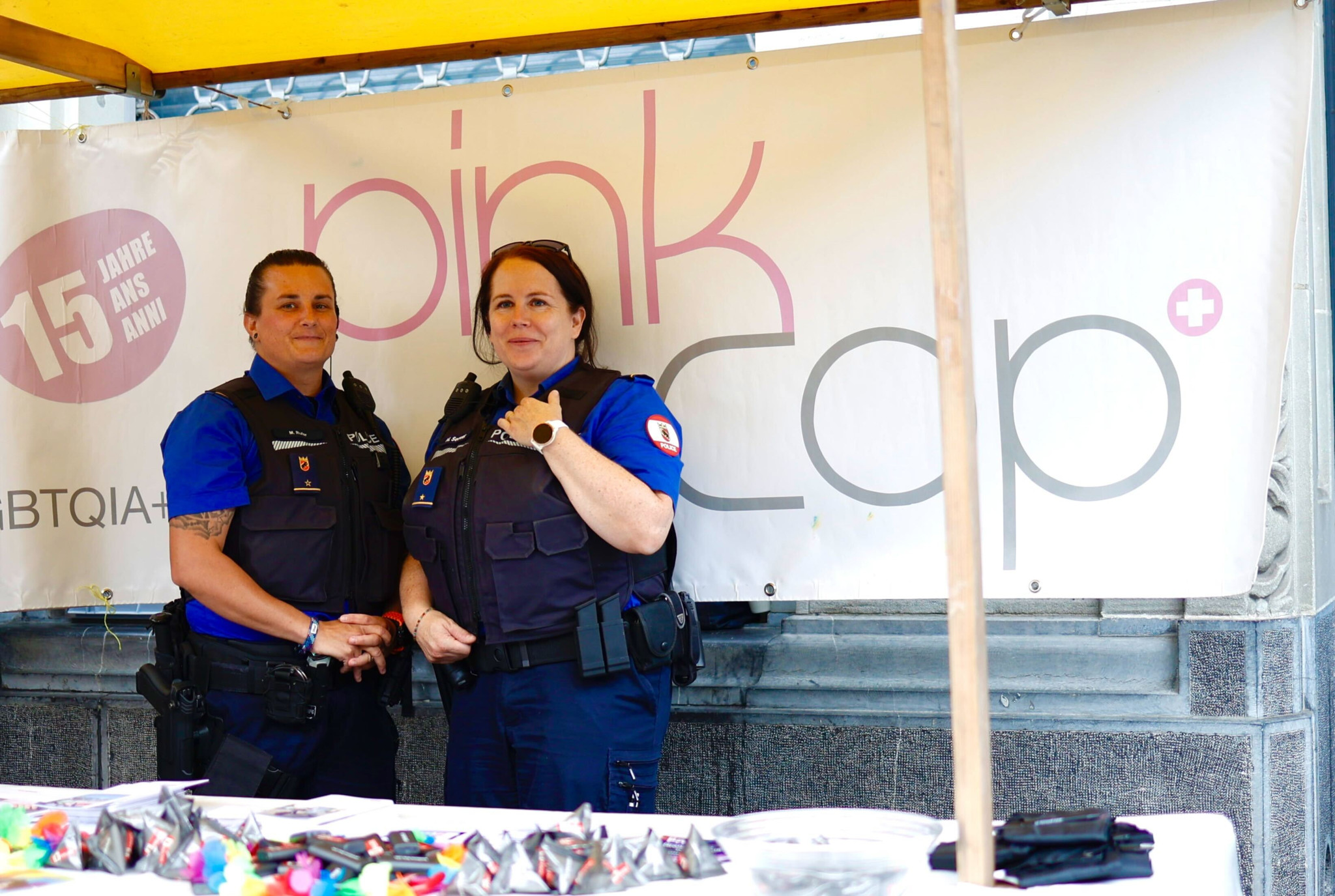 Zwei Polizistinnen in Uniform stehen vor einem Stand mit dem Banner ’pink cop’, der für 15 Jahre Dienstfeier wirbt. Der Tisch ist mit bunten Gegenständen dekoriert. Zwei Polizistinnen in Uniform stehen vor einem Stand mit dem Banner ’pink cop’, der für 15 Jahre Dienstfeier wirbt. Der Tisch ist mit bunten Gegenständen dekoriert.