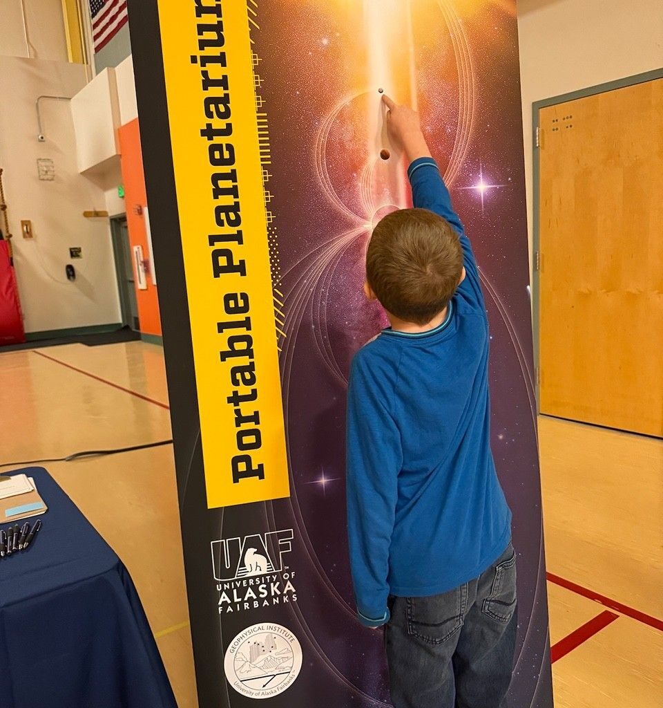 Photo of a child pointing to a planetary body on a banner called "Portable Planetarium" that displays the Sun, planets, magnetic field lines, and stars in the background.