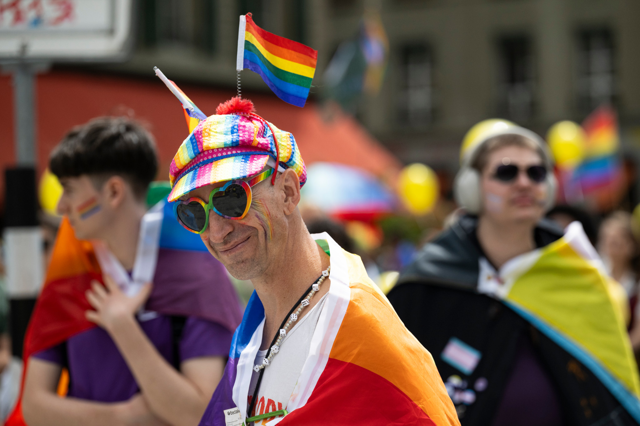 Teilnehmer der BernPride 2025 trägt Regenbogenkleidung und Hut mit Flaggen, lächelnd mit Herz-Sonnenbrille. Teilnehmer der BernPride 2025 trägt Regenbogenkleidung und Hut mit Flaggen, lächelnd mit Herz-Sonnenbrille.