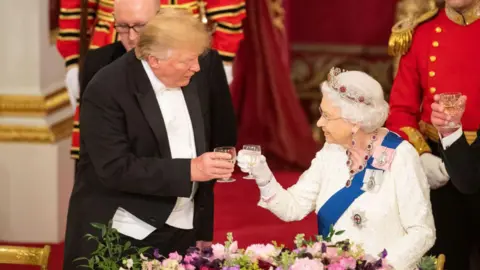 Getty Images Donald Trump raises a glass with Queen Elizabeth II during a state banquet as part of his 2019 visit to the UK. He wears a formal black suit, white shirt and bow tie, while she wears a white, long-sleeved dress, silver crown and a ruby and diamond necklace. The pair are touching wine glasses and appear to be attended by two footmen.