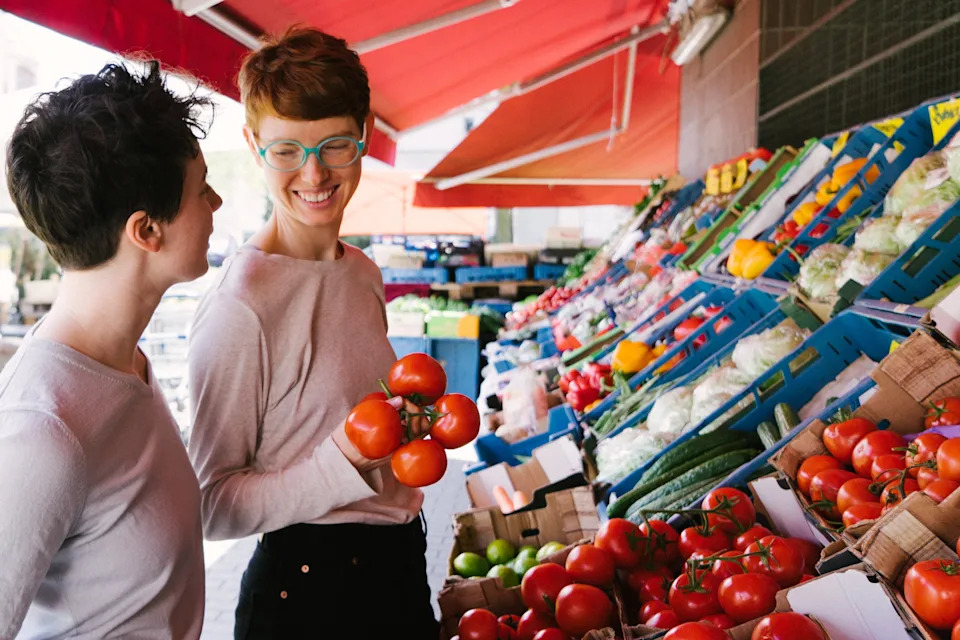 Two people shopping at an outdoor market, smiling and holding tomatoes, with a variety of fresh produce on display in the background