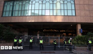 Police officers in high-visibility jackets stand in a line behind metal barriers outside the International Hotel in east London, with the building’s large arched windows visible above the entrance.