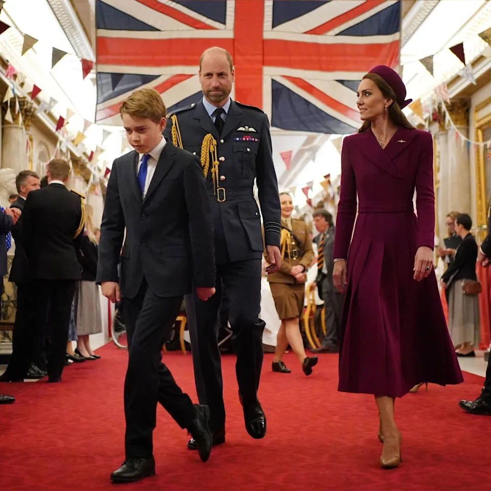 Prince George walking in front of Prince William in a military uniform and Prince Kate in a burgundy coat with a Union Jack behind them.