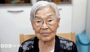 An old woman white short white hair and wire frame glasses sits in a chair and looks into the camera