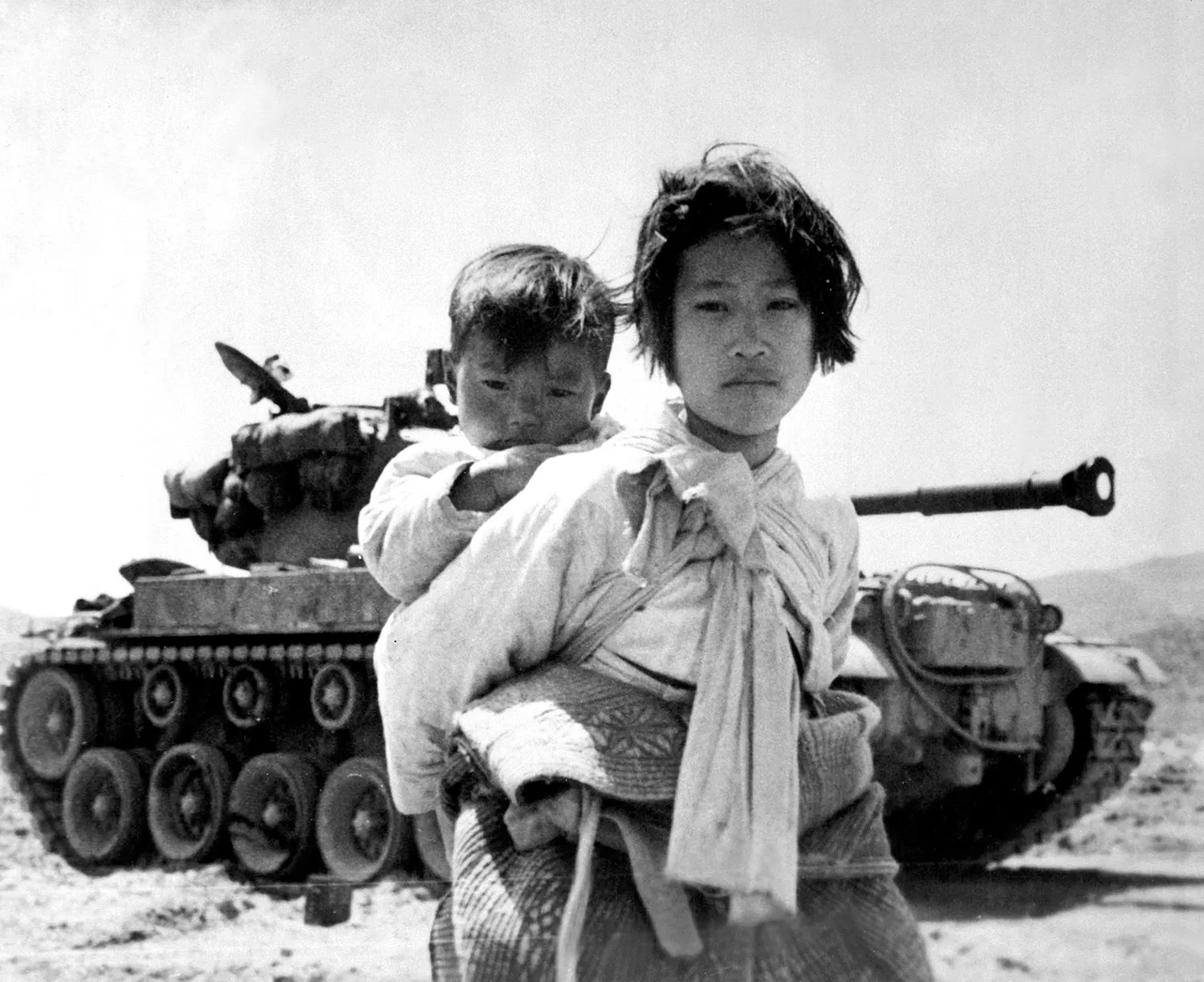 A teenaged girl carries a child on her back in front of a tank.