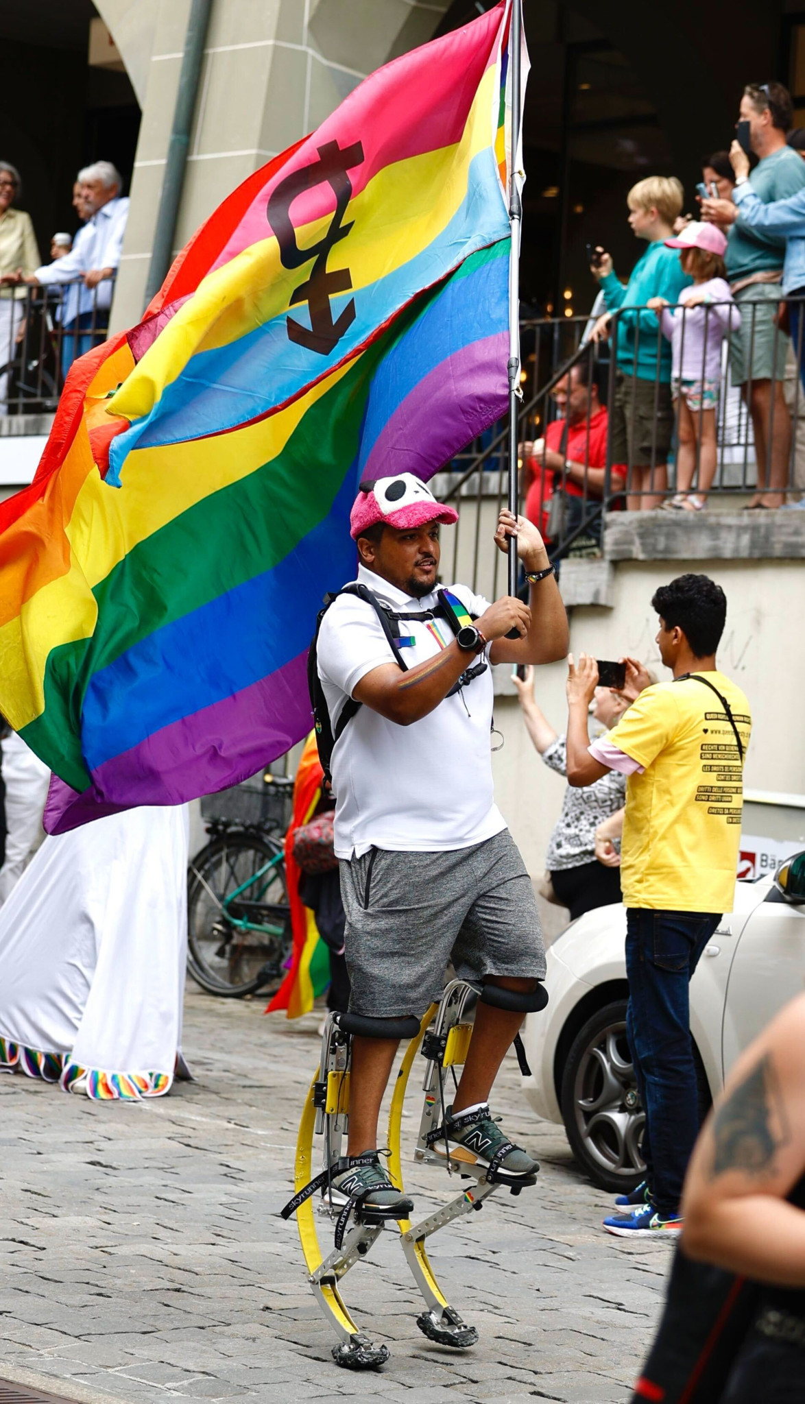 Ein Mann auf Sprungstelzen hält eine Regenbogenflagge mit einem Transgender-Symbol bei einer Parade. Ein Mann auf Sprungstelzen hält eine Regenbogenflagge mit einem Transgender-Symbol bei einer Parade.