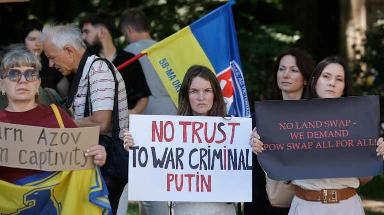 Three women stand holding placards with texts such as "No Trust to War Criminal Putin" with other people, trees and a Ukrainian flag behind them.
