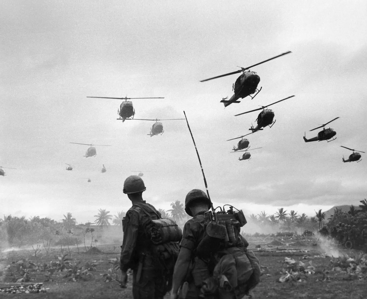 Soldiers in gear and helmets look up toward a sky filled with helicopters. Palm trees are on the horizon in the background.