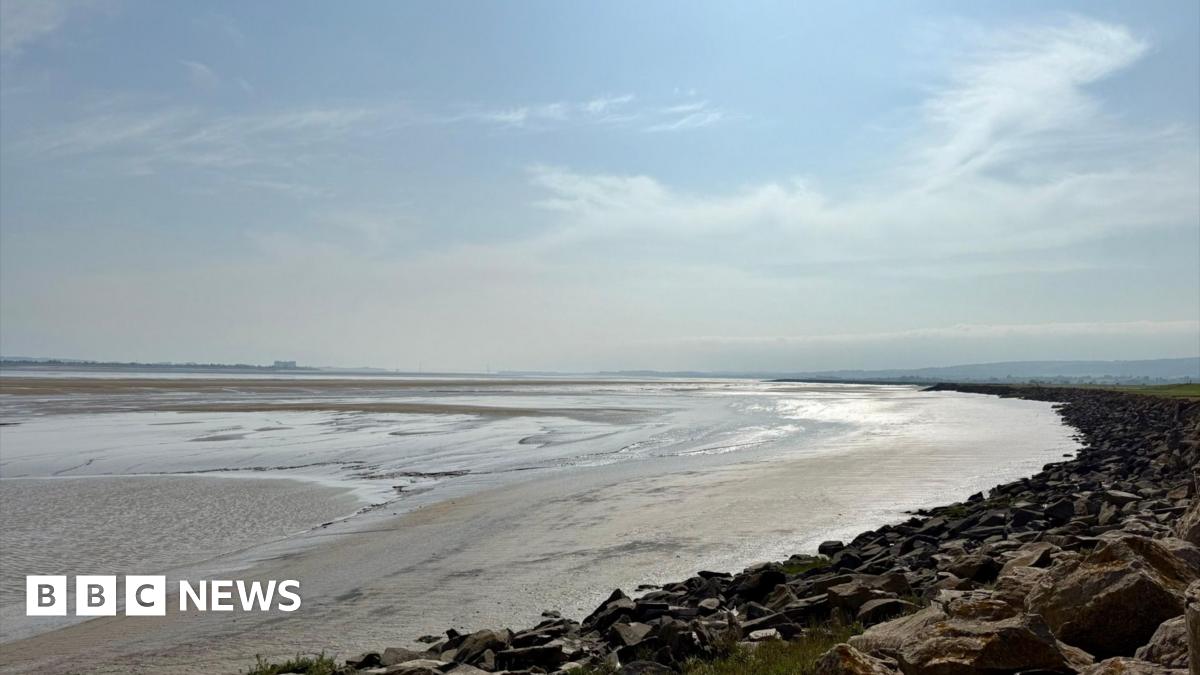 A sweeping coastline stretches out in a semi circle, with the sea so far out it is hardly visible. Large rocks go as far as the eye can see in the right of the photo, with the rocks then meeting the golden sand. The sand appears to be damp with the sunlight shimmering off of it and there is a small pool of water in the right of the image. The sky is blue with a few small clouds.