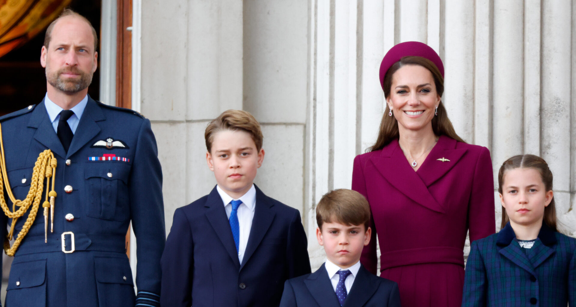 Prince William wears military uniform and Kate Middleton wears a dark red coat dress and matching hat as they stand with children George, Charlotte, and Louis on the Buckingham Palace balcony
