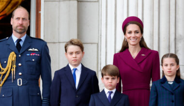 Prince William wears military uniform and Kate Middleton wears a dark red coat dress and matching hat as they stand with children George, Charlotte, and Louis on the Buckingham Palace balcony