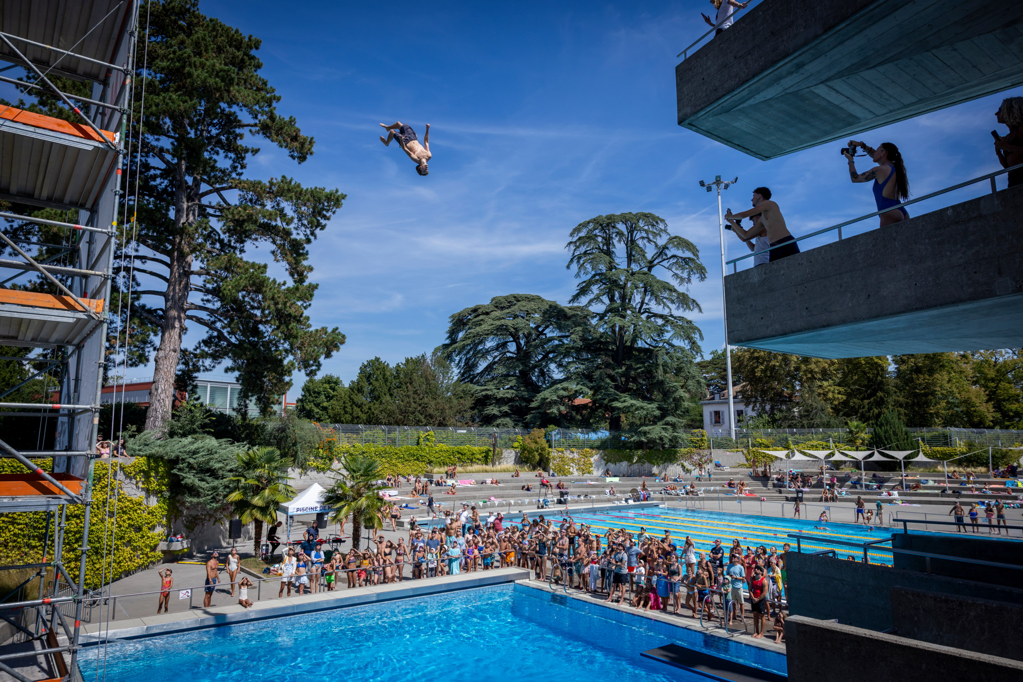 Un plongeur en plein saut lors de l’événement Lancy Cliff Diving à la piscine de Marignac à Genève, avec des spectateurs et des photographes observant.