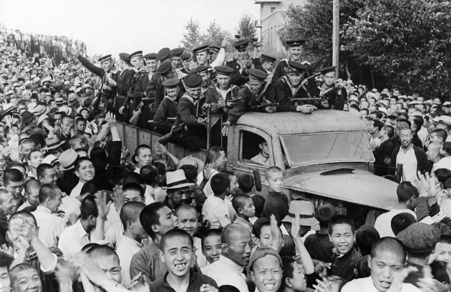 A crowd of people smile and celebrate as a truck carrying sailors drives among them.