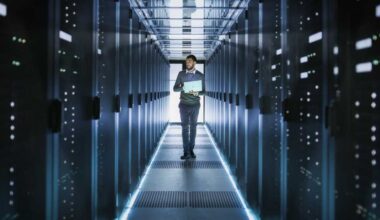 IT Technician Works on Laptop in Big Data Center full of Rack Servers. He Runs Diagnostics and Maintenance, Sets System Up.
