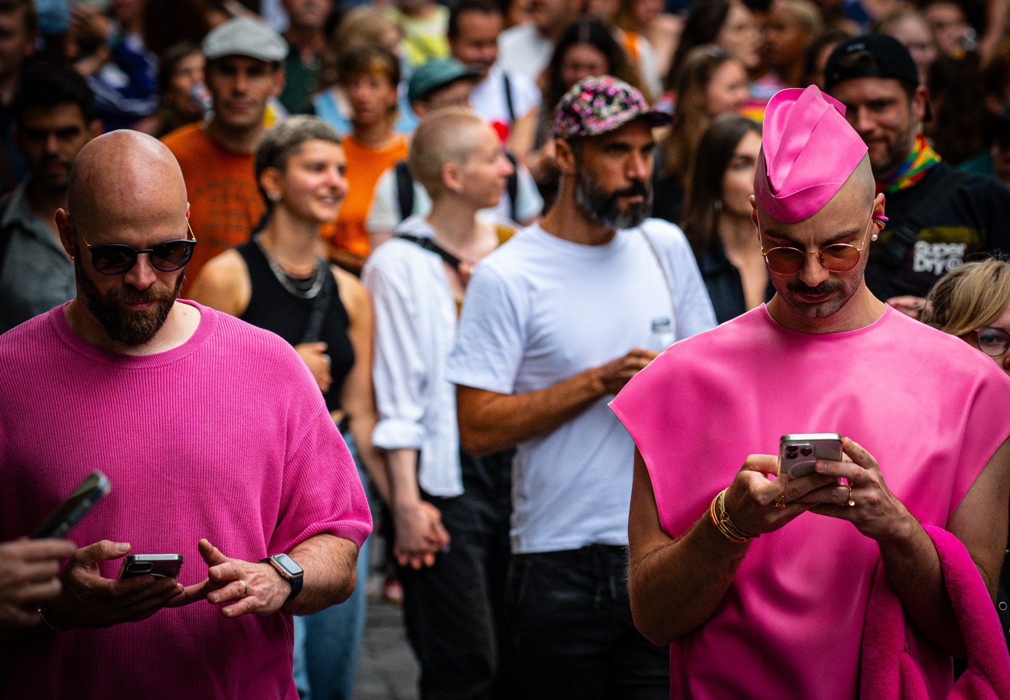 Zwei Männer in auffälligem, pinkem Outfit stehen in einer Menschenmenge und schauen auf ihre Handys. Zwei Männer in auffälligem, pinkem Outfit stehen in einer Menschenmenge und schauen auf ihre Handys.