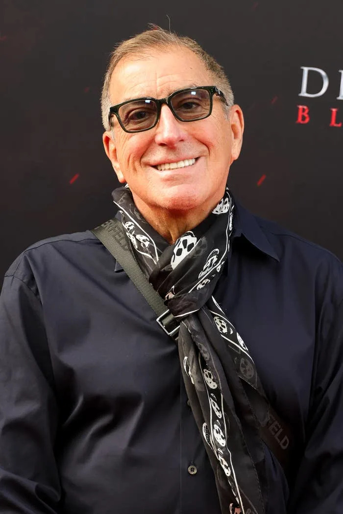 Kenny Ortega in sunglasses smiles on the red carpet, wearing a dark shirt and a scarf with a skull pattern
