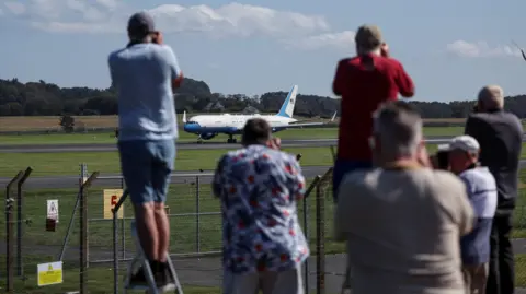 Reuters Plane spotters taking pictures of Air Force Two at Prestwick Airport. Two people, one on the left wearing a light blue top and one on the right wearing a red top, are on step ladders. A man in a multi-coloured white, blue and red top is in the middle of the image. They are blurred. The plane in the background is in full focus and is mainly white with blue accents.
