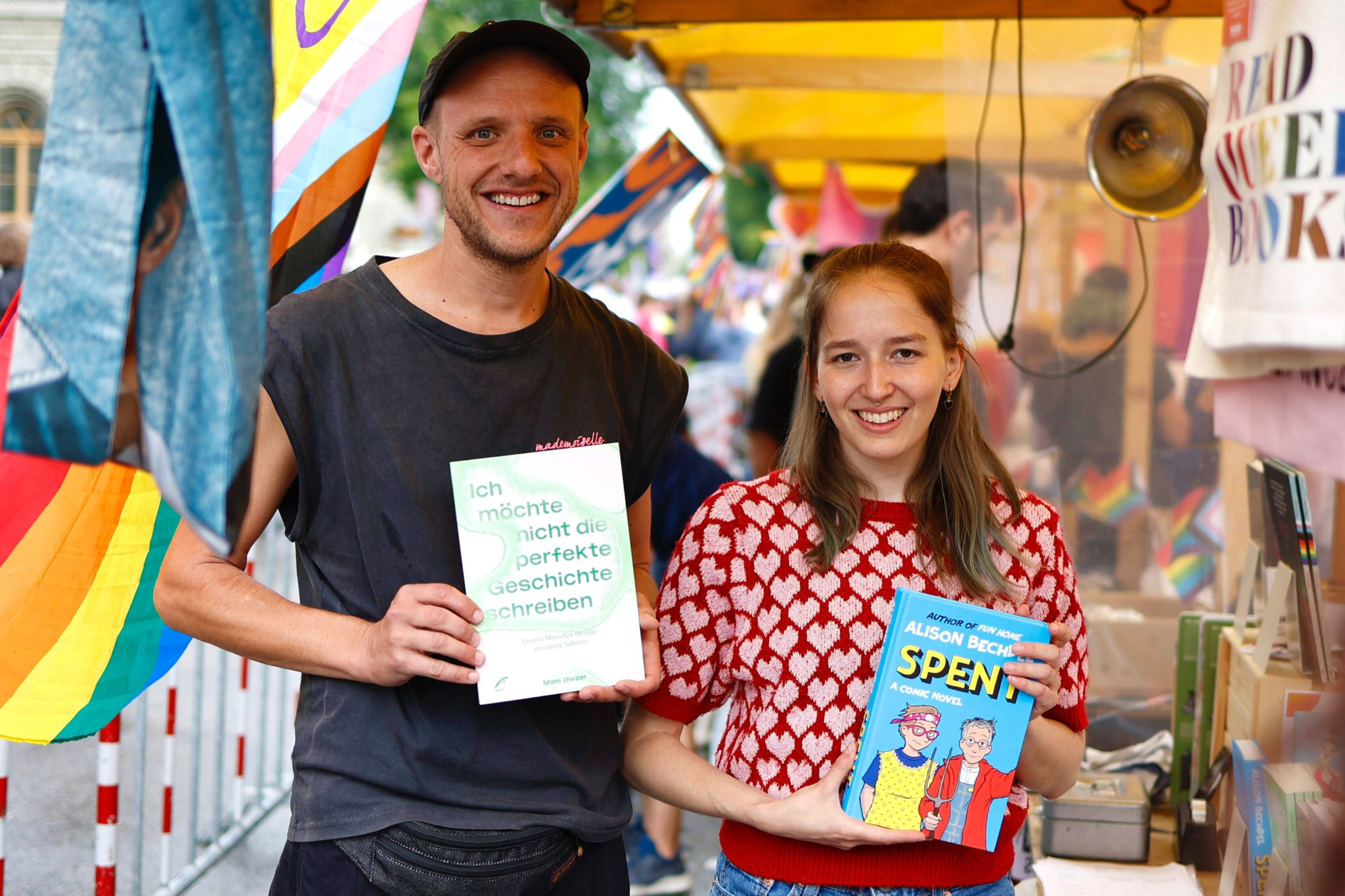 Ein Mann und eine Frau auf einer Buchmesse halten Bücher in den Händen. Die Frau trägt ein rot-weisses Oberteil und hält ein Buch mit dem Titel ’Spent’. Der Mann trägt ein schwarzes Oberteil und eine Mütze, hält ein Buch mit grünem Text. Im Hintergrund sind bunte Fahnen und ein gelber Pavillon zu sehen. Ein Mann und eine Frau auf einer Buchmesse halten Bücher in den Händen. Die Frau trägt ein rot-weisses Oberteil und hält ein Buch mit dem Titel ’Spent’. Der Mann trägt ein schwarzes Oberteil und eine Mütze, hält ein Buch mit grünem Text. Im Hintergrund sind bunte Fahnen und ein gelber Pavillon zu sehen.