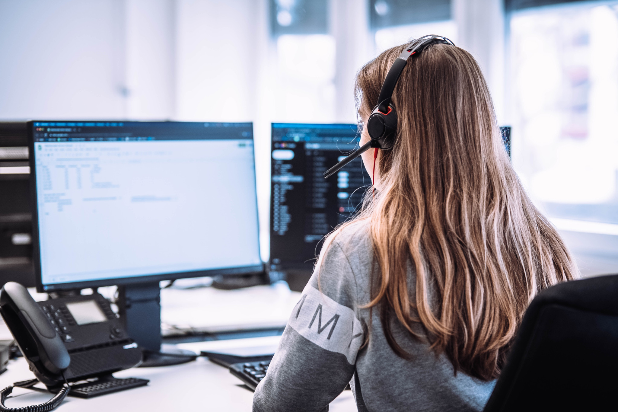 Eine Callcenter-Agentin mit Headset sitzt vor mehreren Computerbildschirmen. Sie trägt einen grauen Pullover. Eine Callcenter-Agentin mit Headset sitzt vor mehreren Computerbildschirmen. Sie trägt einen grauen Pullover.
