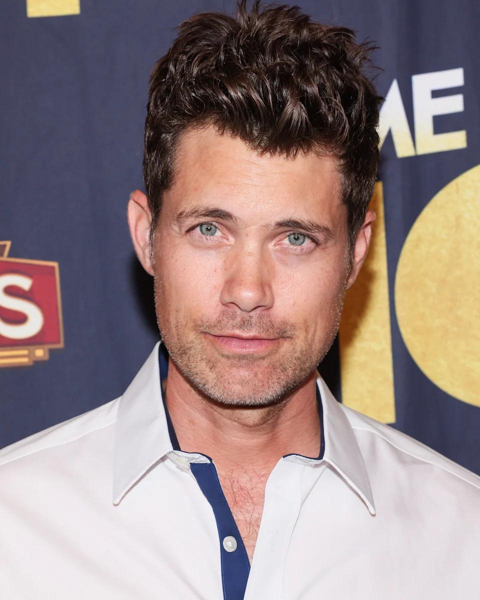 Drew Seeley with short, styled hair wearing a white shirt with a dark collar stands against a backdrop, likely at a red carpet event