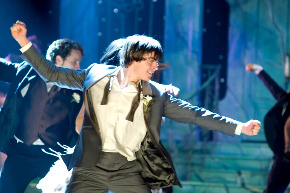 Zac Efron in a suit, shirt untucked, dancing energetically at a formal event with others in the background