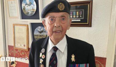 Tommy Doherty  stands in a room in his home, in front of a wall on which medals and a number of plates displaying images of the war are scene. Tommy is wearing a war blazer and beret, and a shirt and tie. He wears a number of military badges and medals on his blazer.