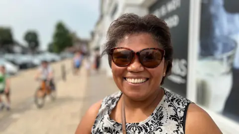 A lady smiling at the camera wearing big sunglasses. She is standing on a high street with a cyclist passing who is out of focus.