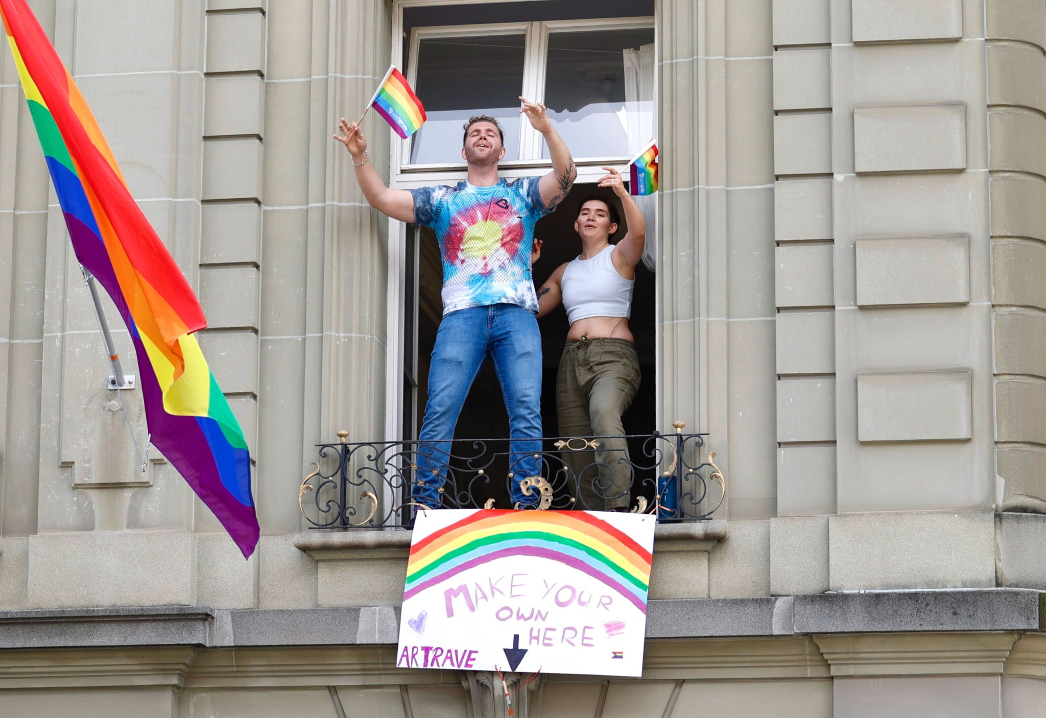 Zwei Personen stehen in einer offenen Fensteröffnung eines Gebäudes und schwenken bunte Regenbogenflaggen. Ein Schild mit der Aufschrift ’Make Your Own Here’ ist unter dem Fenster angebracht. Zwei Personen stehen in einer offenen Fensteröffnung eines Gebäudes und schwenken bunte Regenbogenflaggen. Ein Schild mit der Aufschrift ’Make Your Own Here’ ist unter dem Fenster angebracht.