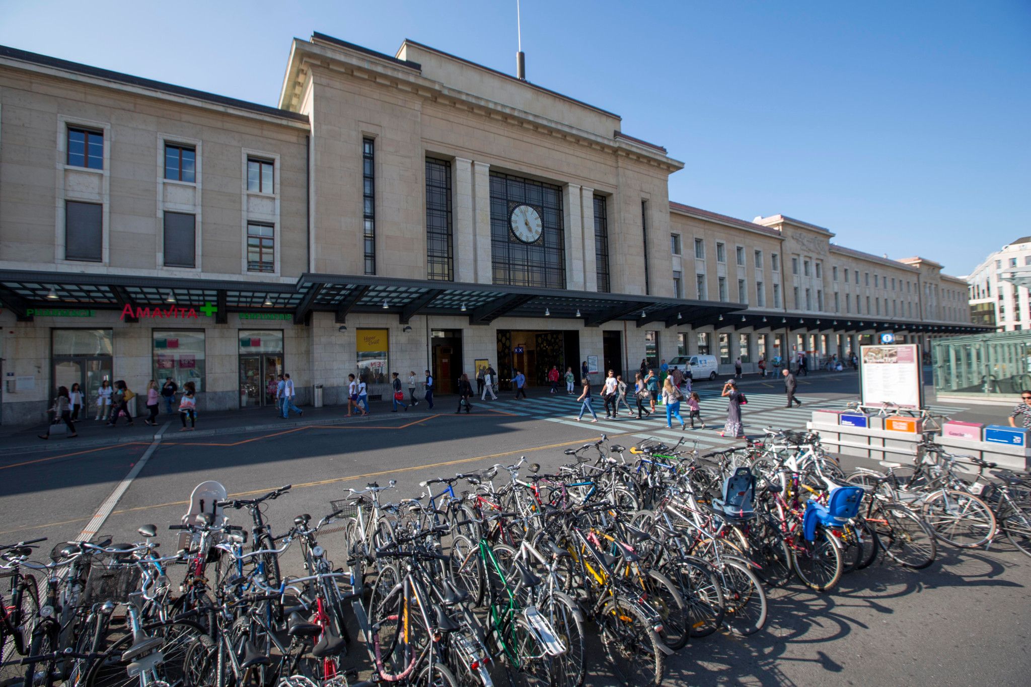 Façade de la gare de Cornavin à Genève avec de nombreux vélos garés devant et des passants marchant à l’entrée sous un ciel ensoleillé.