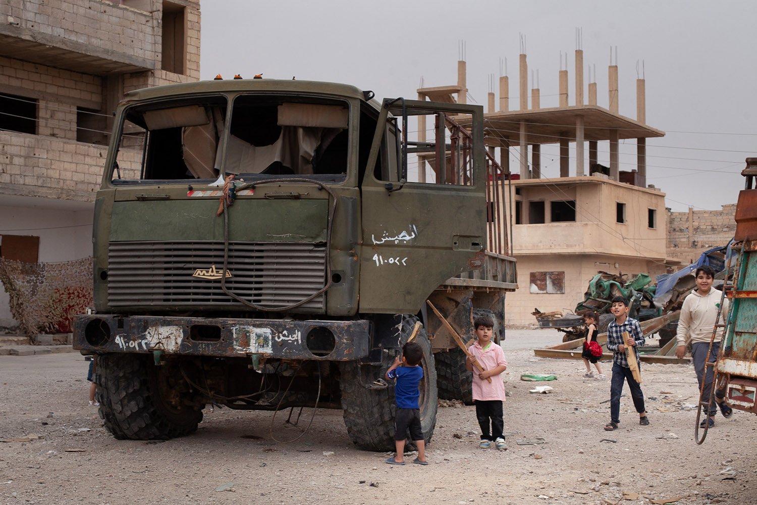 Five children, some holding sticks, play around a military vehicle with its door ajar. Half constructed and damaged buildings in the background.