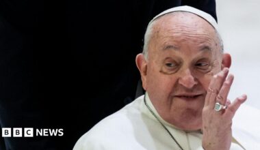 Pope Francis waving his left hand against a black and white background