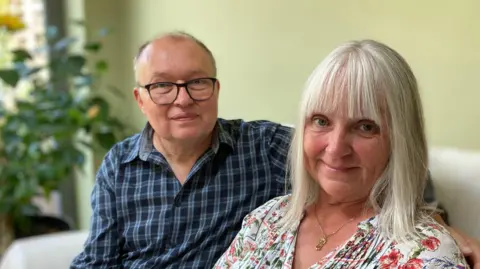 Mr and Mrs Rathgay are sitting on a cream sofa with a light green wall behind them. Mr Rathgay on the left is balding, wearing a blue checked shirt and black glasses. Mrs Rathgay on the right has white shoulder-length hair with a fringe, and is wearing a flowery blouse.