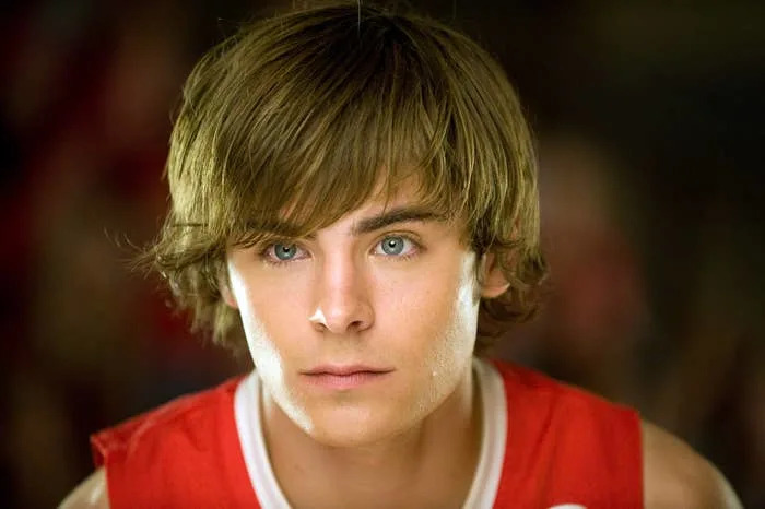 Zac Efron with medium-length hair wearing a casual top looks forward with a focused expression