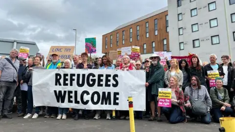 A group of people stand outside a large white and wood-panelled building. They hold a large sign reading "refugees welcome" as well as smaller signs with the same message. Another sign says "love thy neighbour". 