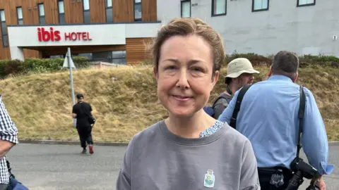 Councillor Chloe Turner looks at the camera. She wears a grey sweatshirt. The sign for the Ibis Hotel is visible in the background.