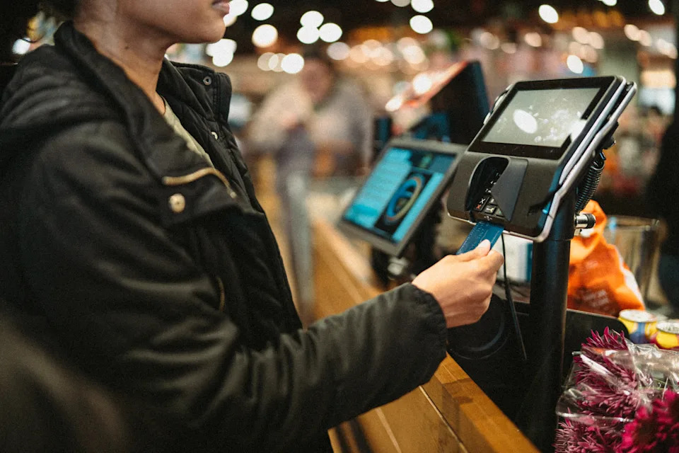 Person swipes card at a self-checkout machine in a busy store, holding a bag of groceries