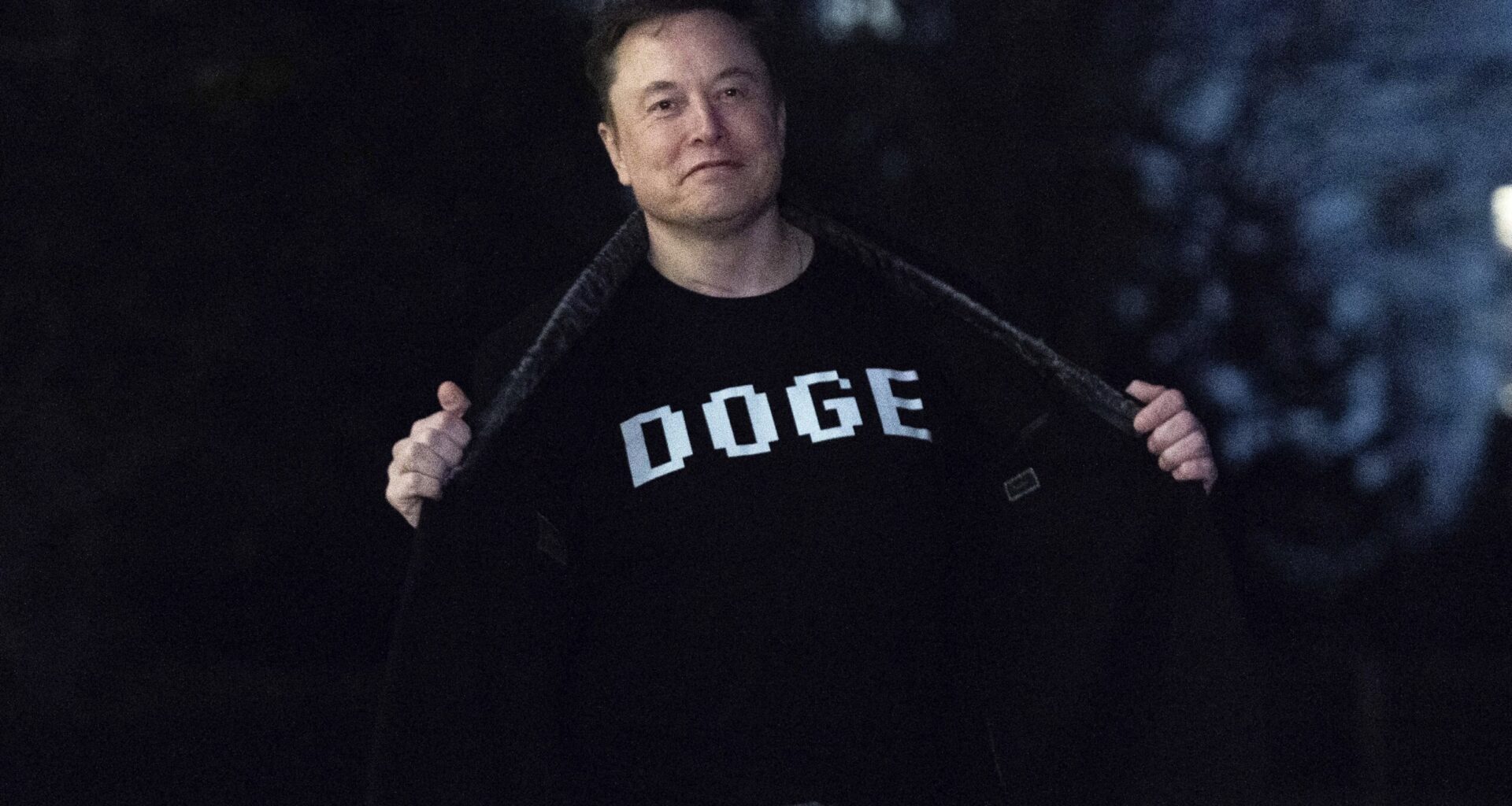 A person stands outdoors at night, opening their jacket to reveal a T-shirt with the word DOGE printed in bold letters.