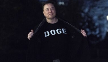 A person stands outdoors at night, opening their jacket to reveal a T-shirt with the word DOGE printed in bold letters.