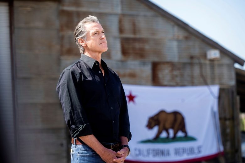 A person stands in front of a rustic building with the California flag in the background.
