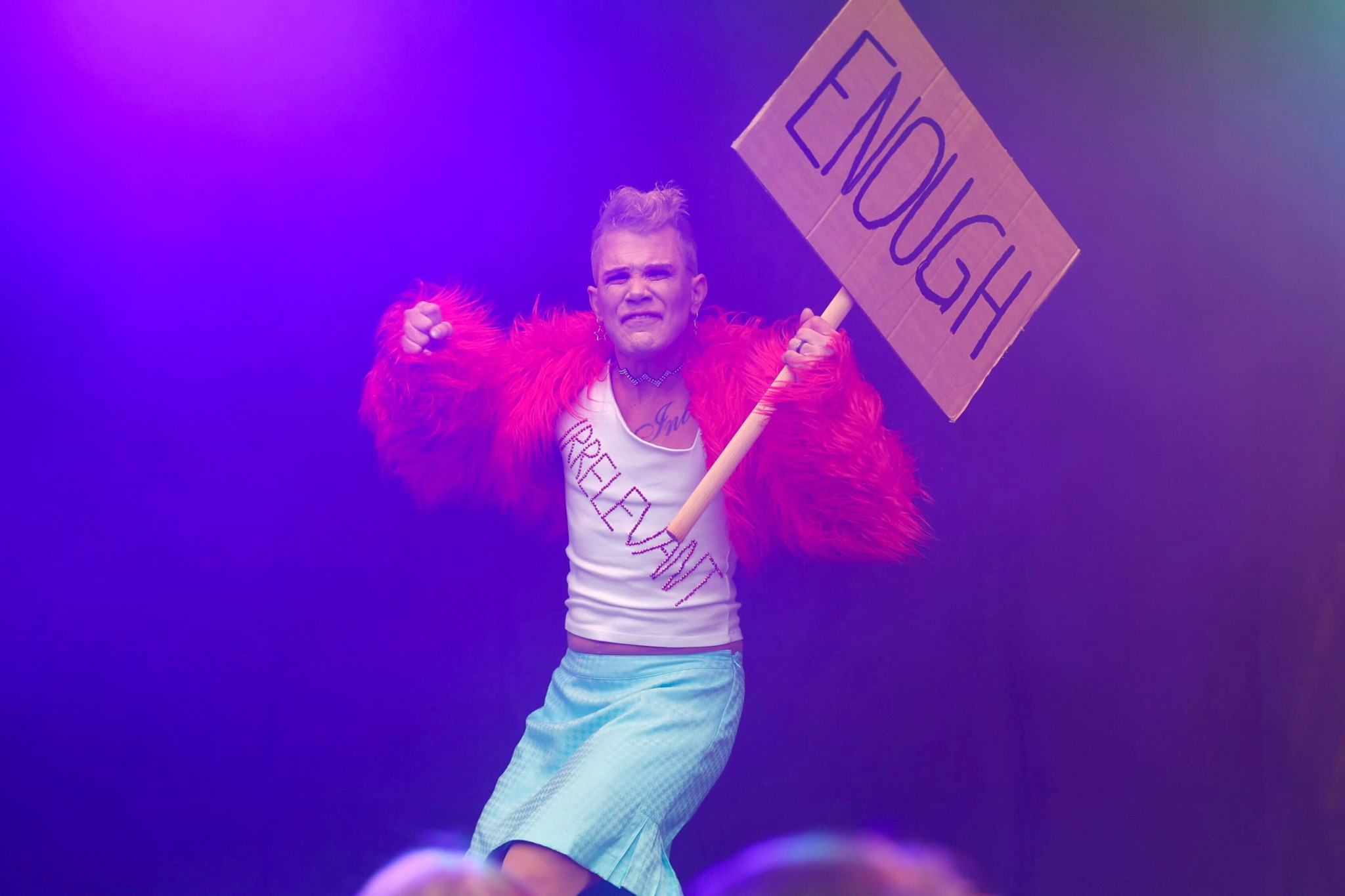 Person in rotem Fellmantel hält Schild mit der Aufschrift ’ENOUGH’ vor lila Hintergrund. Person in rotem Fellmantel hält Schild mit der Aufschrift ’ENOUGH’ vor lila Hintergrund.