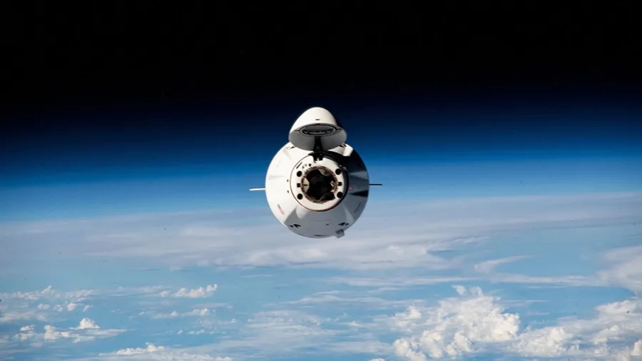 a white space capsule is seen head-on above earth, with cloudy blue skies in the background