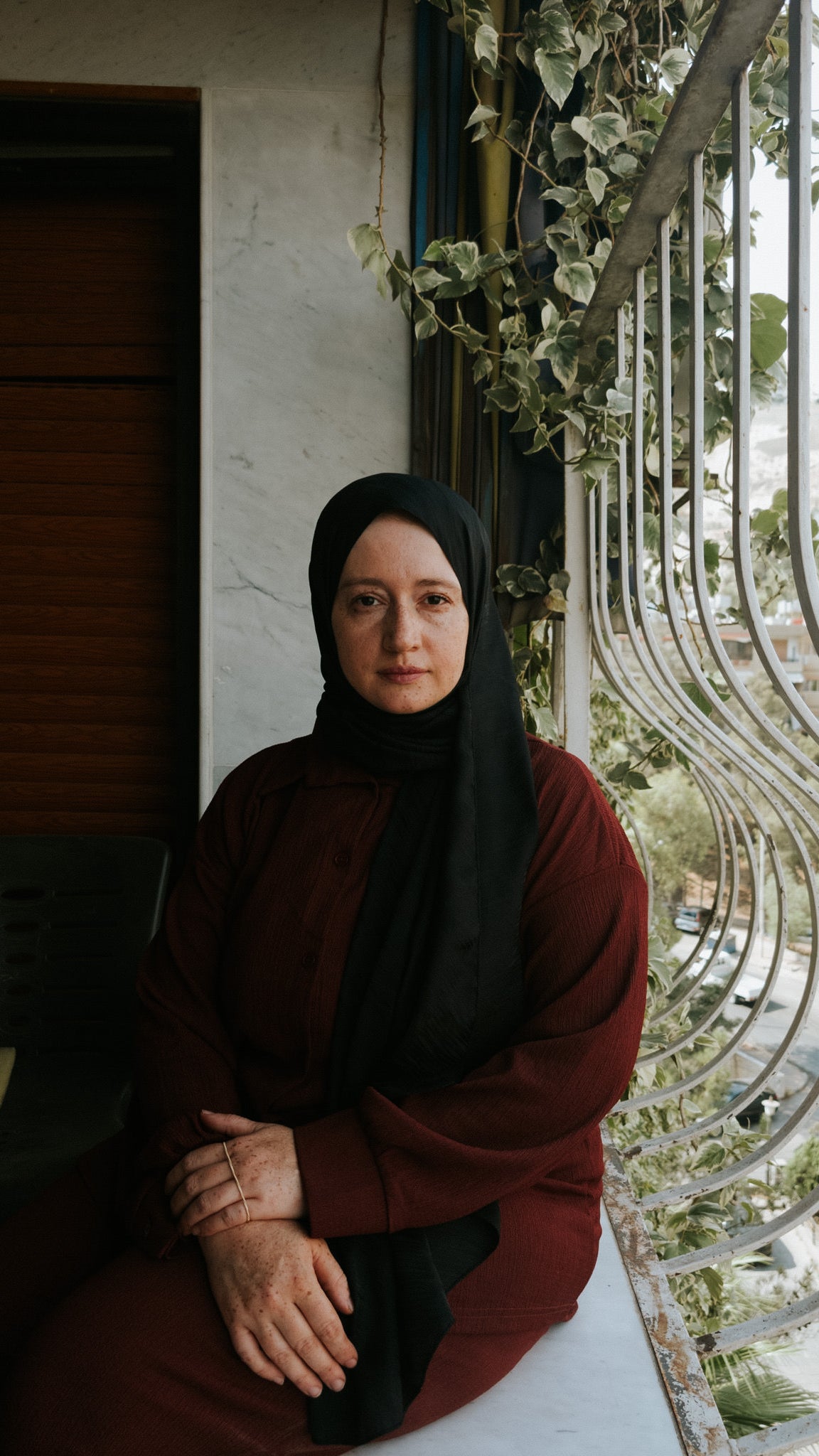 A woman in a black hijab and a burgundy dress sits by a window with metal bars, surrounded by greenery. She has a calm expression and her hands are clasped in front of her. The background reveals a view of trees and buildings outside.
