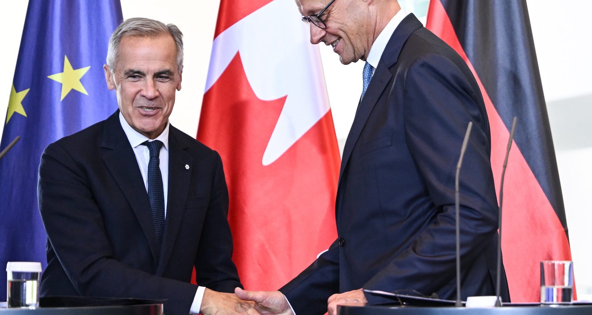 Mark Carney shaking hands with German Chancellor Friedrich Merz, with EU, Canadian and German flags in the background. Both men are smiling.
