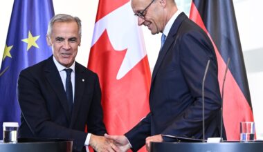 Mark Carney shaking hands with German Chancellor Friedrich Merz, with EU, Canadian and German flags in the background. Both men are smiling.