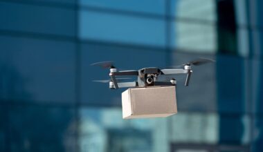 A drone in mid-air with cuboidal cardboard box slung beneath, presumably a postal package, against a background of a building, the blurred rectlinear shapes of windows fill the image, in which appears to be reflected some light-coloured buildings in sunlight