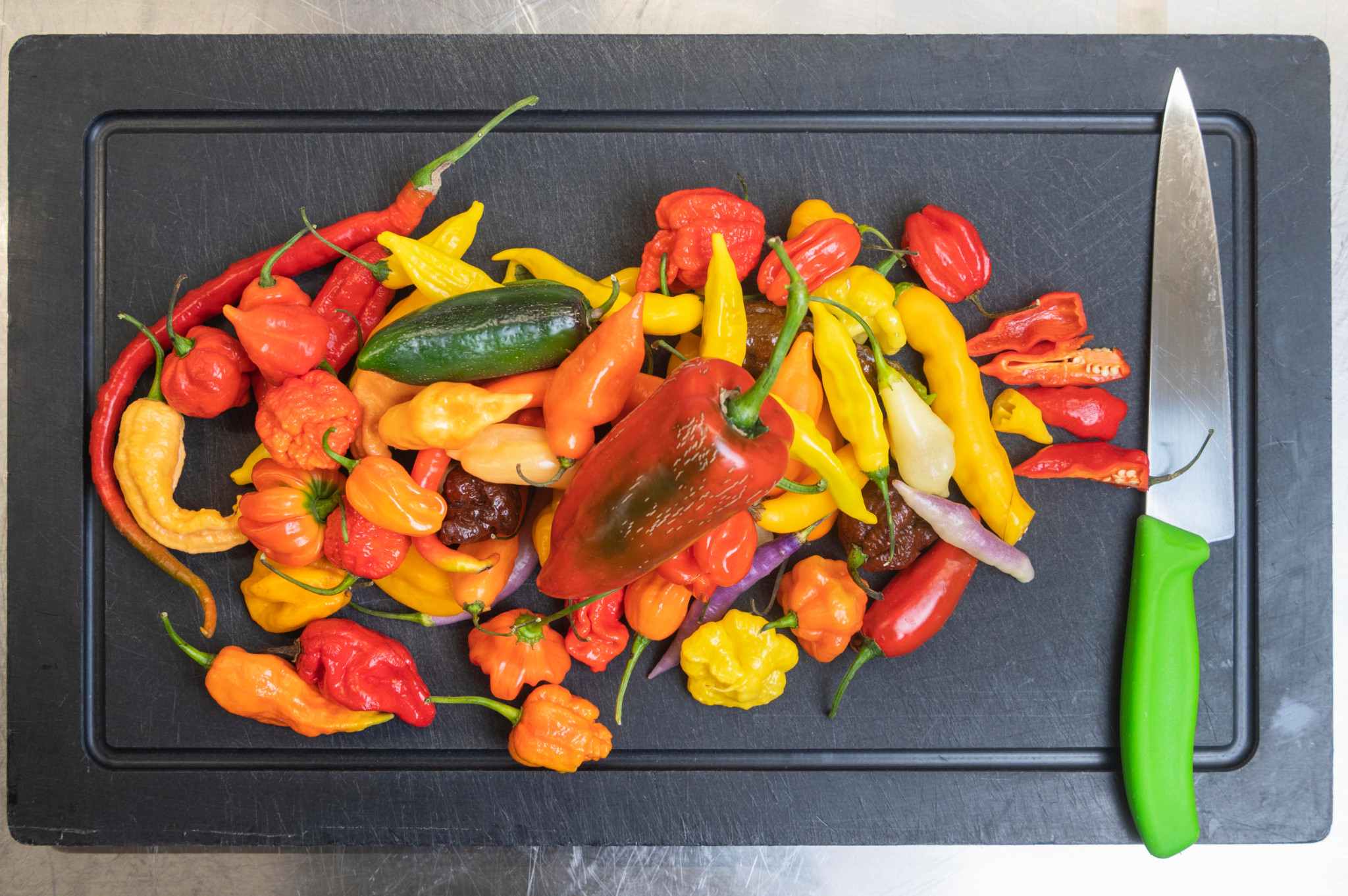 Assortiment de piments colorés sur une planche à découper noire avec un couteau vert, produits par Lucas Malacari de Pica & Repica.