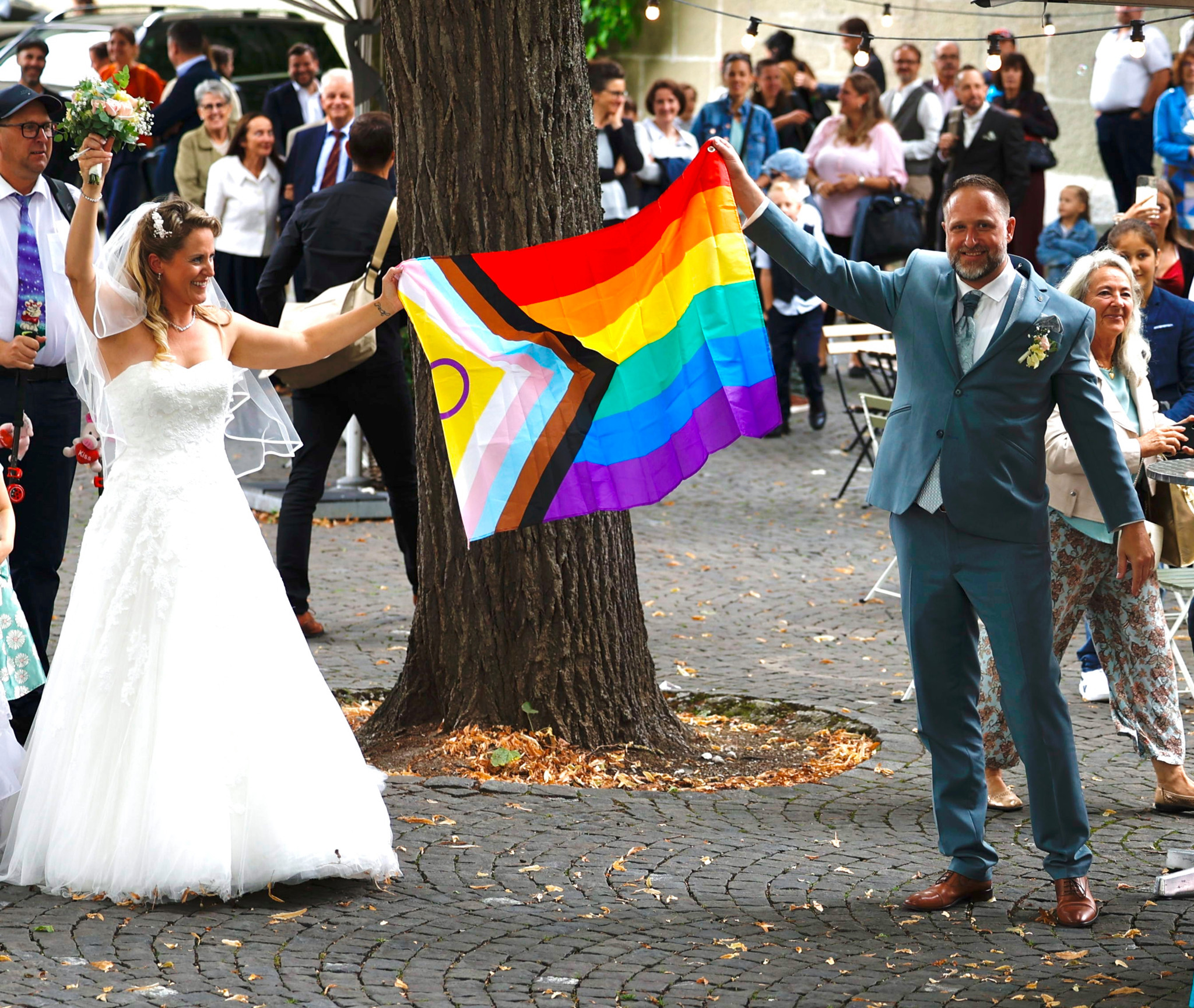 Ein Brautpaar in Hochzeitskleidung hält gemeinsam eine Regenbogen-Pride-Flagge hoch, umgeben von applaudierenden Gästen. Ein Brautpaar in Hochzeitskleidung hält gemeinsam eine Regenbogen-Pride-Flagge hoch, umgeben von applaudierenden Gästen.