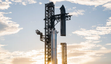 a giant silver spacecraft is stacked atop its silver booster by a metal launch tower