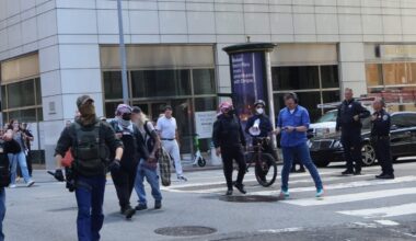 A group of people, some wearing tactical gear and masks, walk across a crosswalk in an urban area with tall buildings and parked cars.
