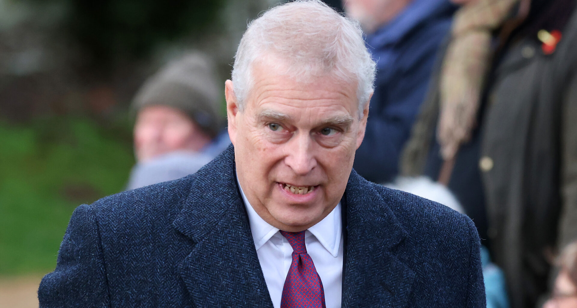 Prince Andrew has white gray hair and wears a large overcoat with a white shirt and red tie underneath